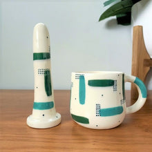 Load image into Gallery viewer, A 5 inch classic ceramic dildo and a matching mug stand on a wooden table. The patterns on the products are teal and forest green strokes, with sets of dark green dots over-layed in sections, with more dots scattered across the white base colour.