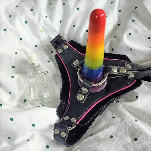A rainbow gradient classic dildo stands in a black harness sitting on a white sheet with dark green dots. A bottle of Uberlube sits to the left.