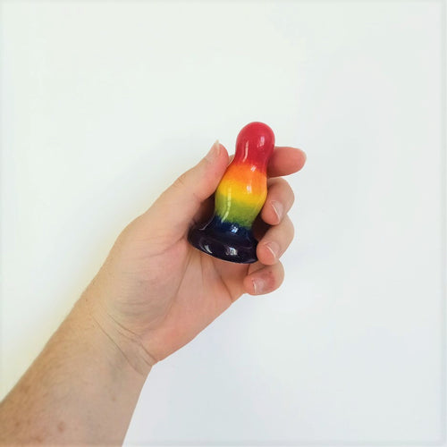 A hand holds a 3 inch ceramic butt plug with 2 bumps in a rainbow gradient pattern in front of a white background.