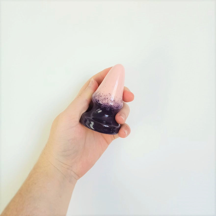 A hand holds a 3 inch cone shaped ceramic butt plug in a dark purple to pink gradient pattern in front of a white wall.