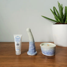 Load image into Gallery viewer, A tube of Bonk organic water-based lubricant, a 6 inch curved ceramic dildo and a matching massage candle in a dark blue to light blue speckle pattern stand in a row on a wooden table. An aloe vera plant in a white pot is visible to the right against a white background.
