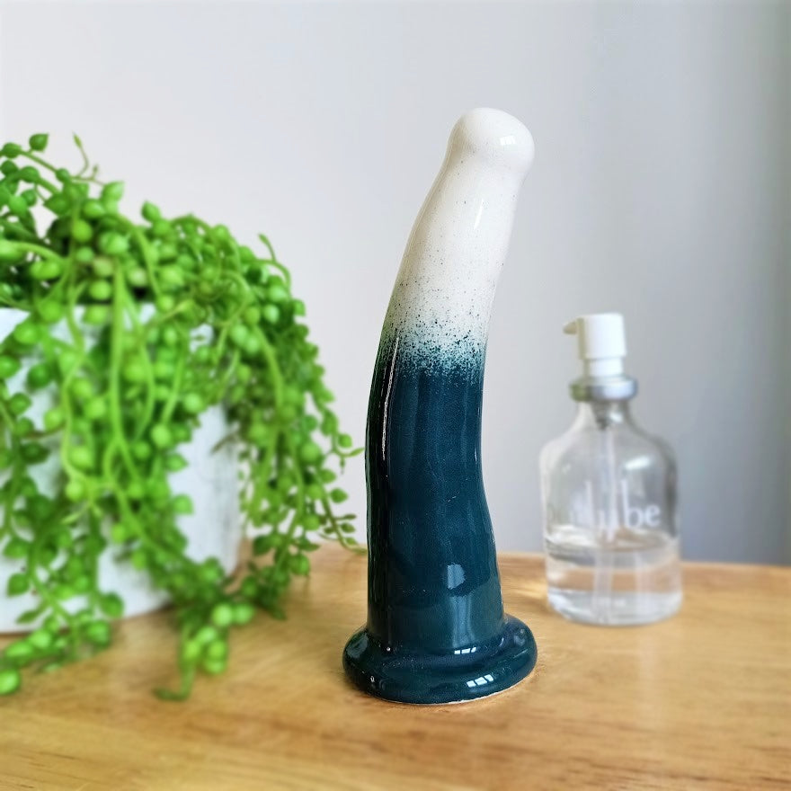 A 6 inch curved ceramic dildo in a white and dark green gradient pattern stands on a wooden table. A glass bottle of Uberlube stands to the right in the background, while a bright green plant flows from a white pot to the left.