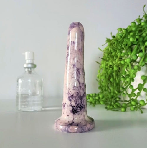 A 4 inch classic ceramic dildo in a pink and dark purple bubble pattern stands on a white table against a white background. A glass bottle of Uberlube is to the left in background, while a bright green pot plant flows from a white pot to the right.