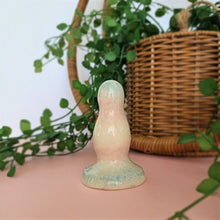 Load image into Gallery viewer, A 3 inch ceramic butt plug with 2 bumps in a trans pride speckle gradient pattern stands on a pink surface with a white background. A green plant flows from a wicker basket behind the butt plug.

