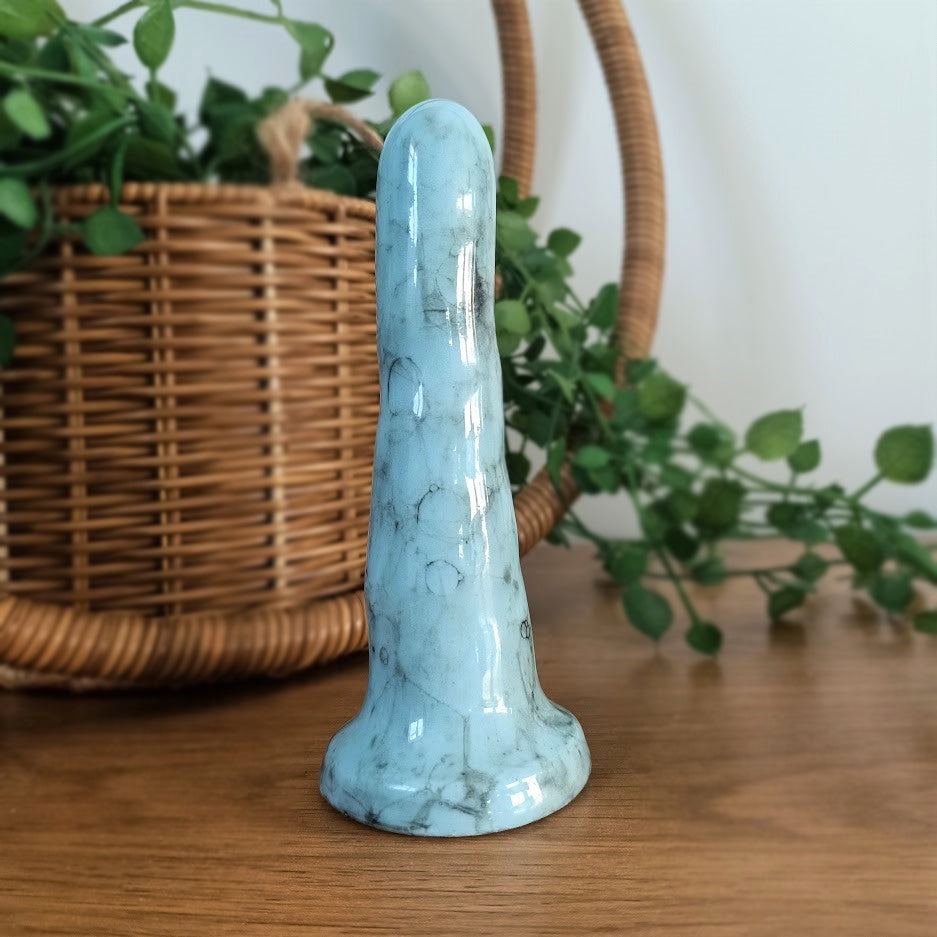 A 5 inch classic ceramic dildo in light blue with a black bubble pattern stands on a wooden surface with a white background. A green plant flows from a wicker basket in the background.